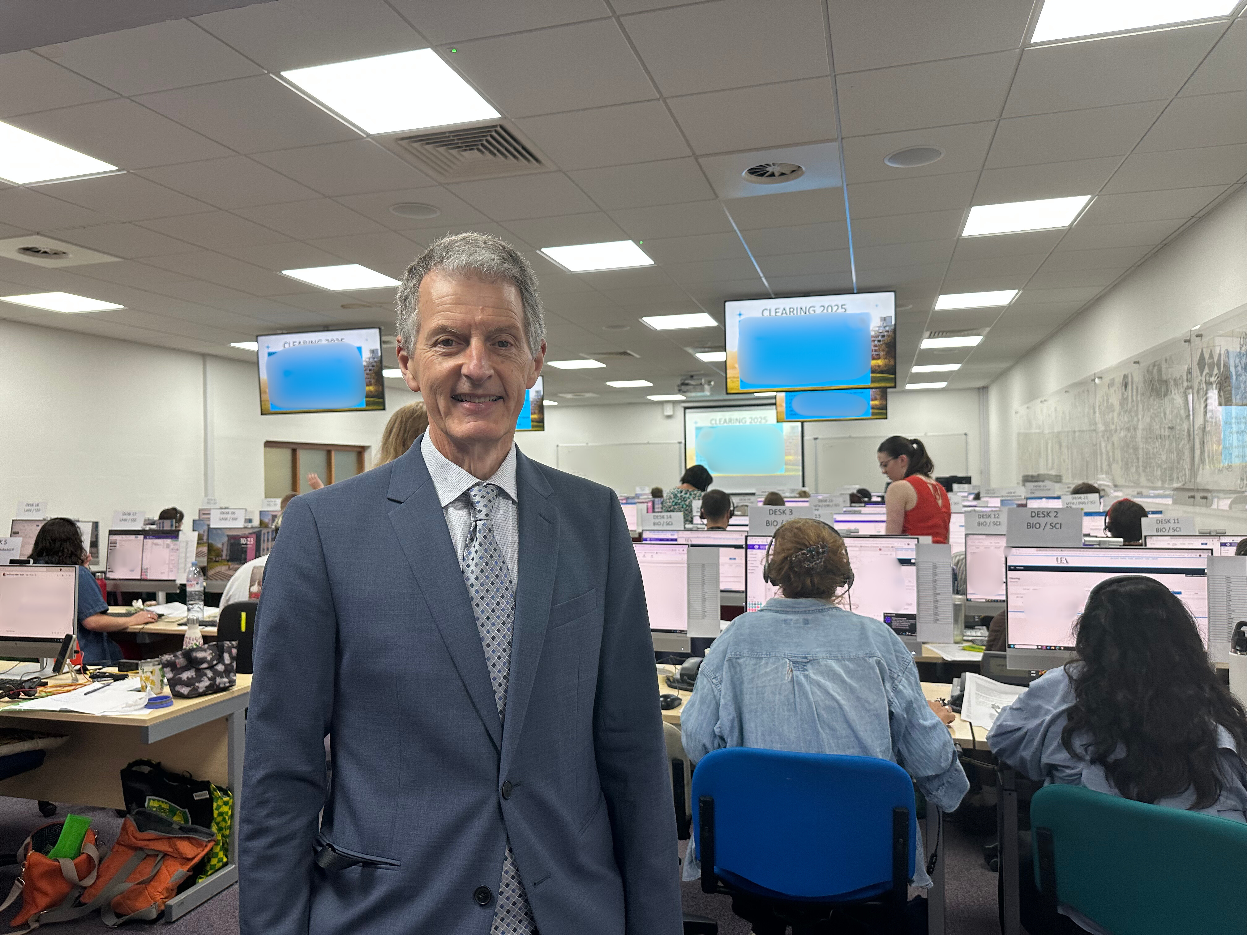 David Maguire stands in front of an office of people sat facing away from the camera on computers. He is smiling, wearing a suit, and on the left side of the picture.