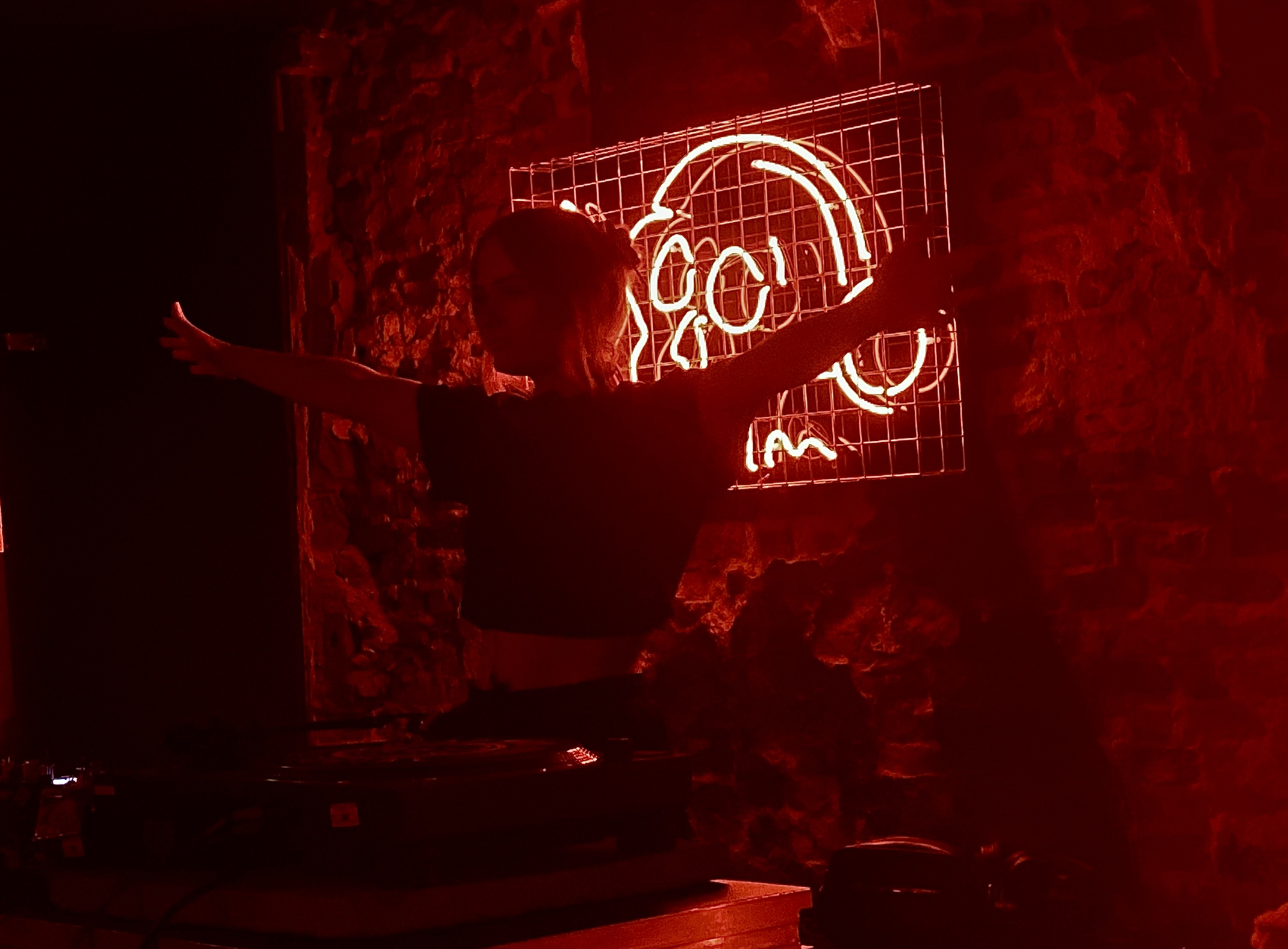 A girl stands with her arms spread out with a DJ deck infront of her. The room is dark, with a red skull wearing headphones lighting the room red.