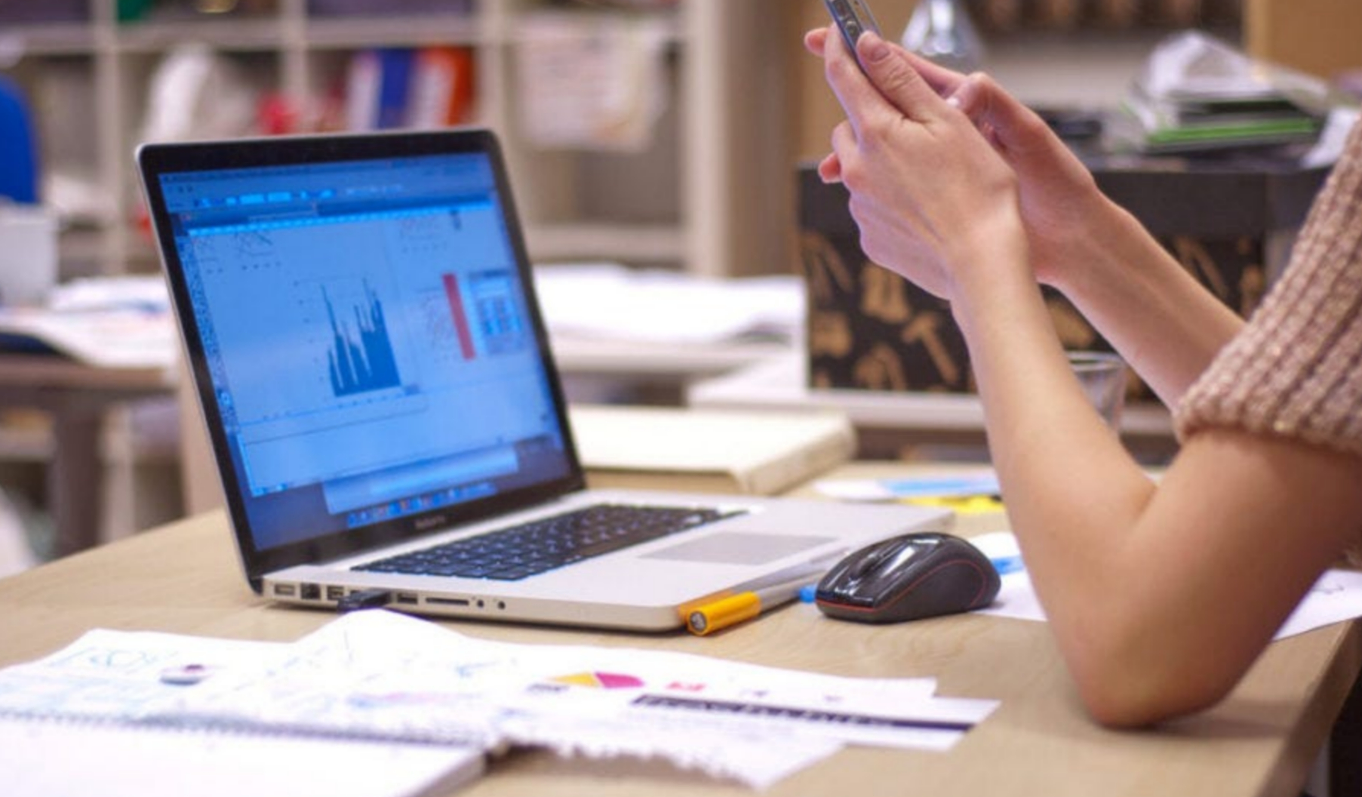 Image of person sat at a desk, with their laptop open. They are sat in front of it, while on their phone.